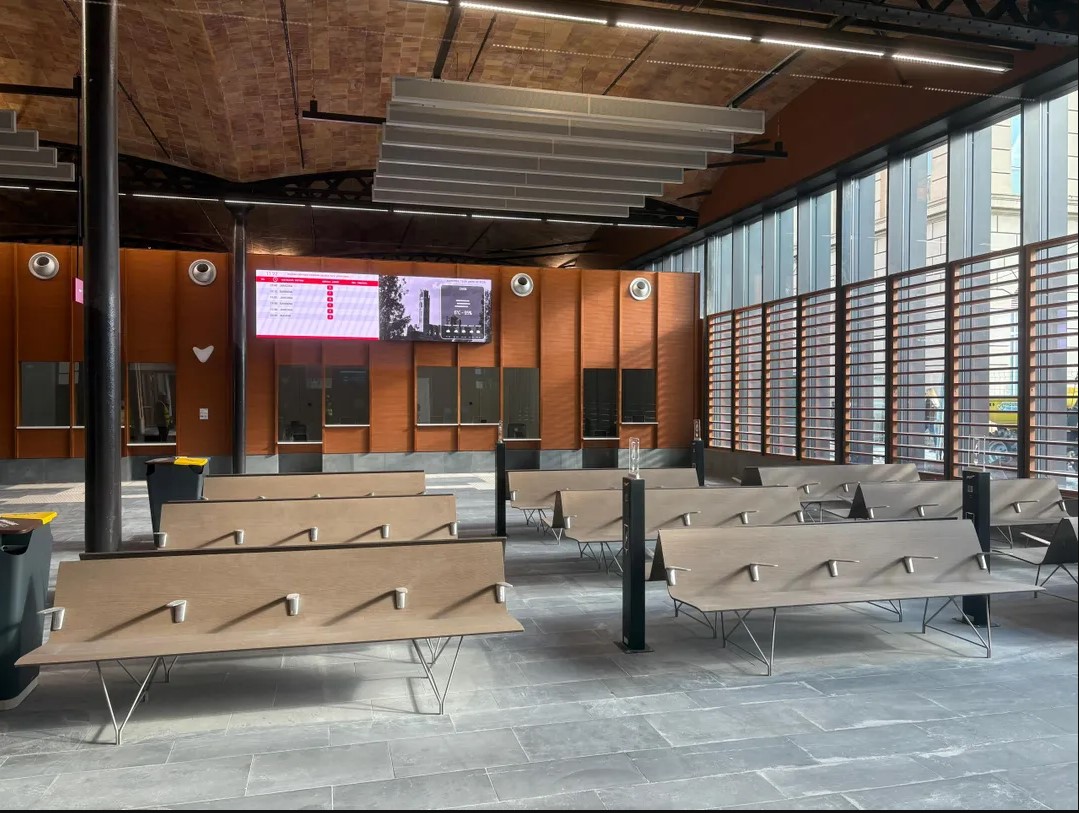 Sala d'espera de l'estació de busos de Lleida amb fileres de bancs de fusta sobre estructura metàl·lica. Al fons, pantalles digitals amb informació de sortides i taquilles. A la dreta, façana acristalada amb llum natural.