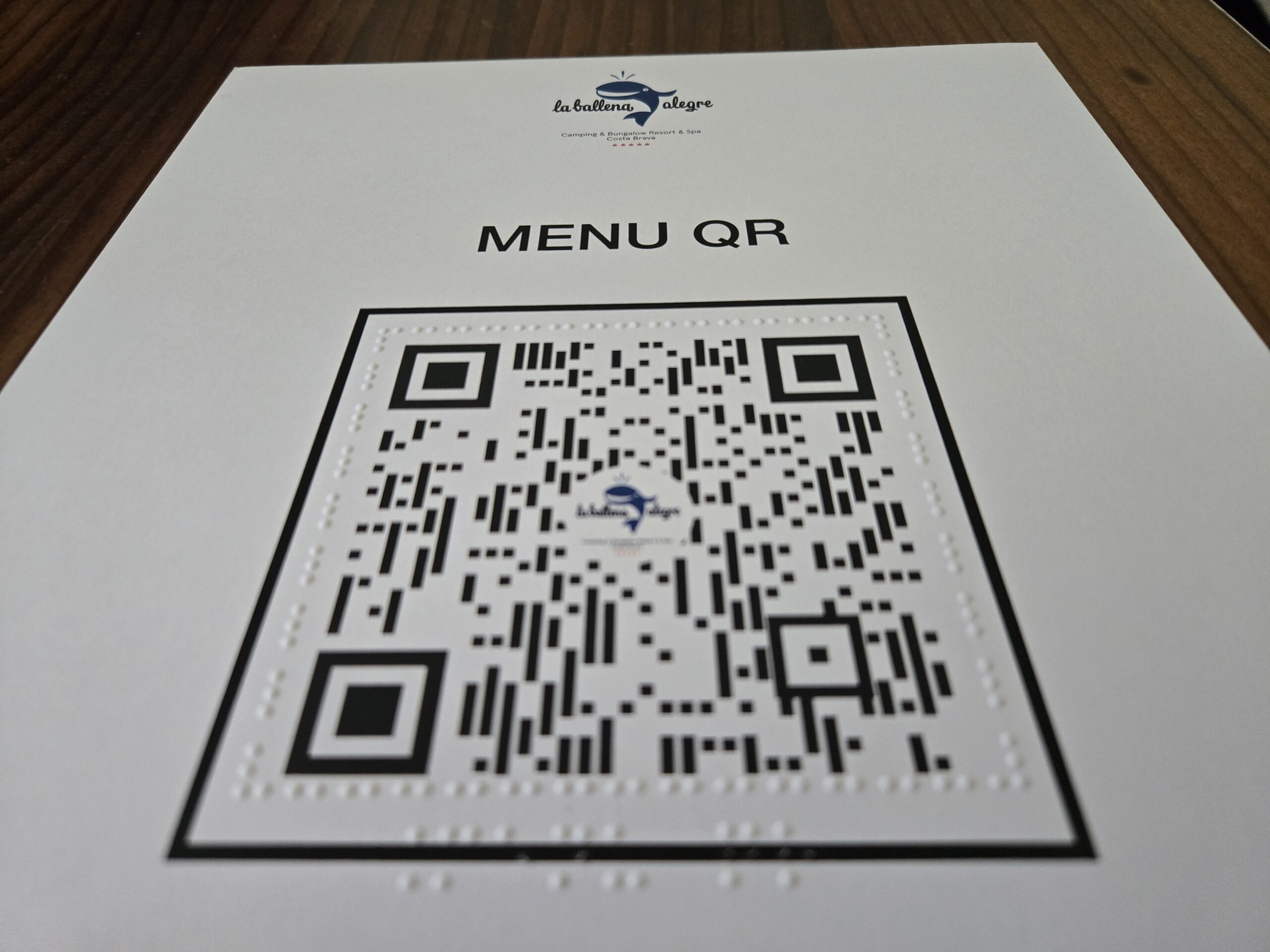 Primer pla d'un codi QR sobre fons blanc, envoltat de punts braille en relleu. Al centre del codi, el logotip de La Ballena Alegre. A la part superior del full, el logotip del càmping i el títol "MENU QR".