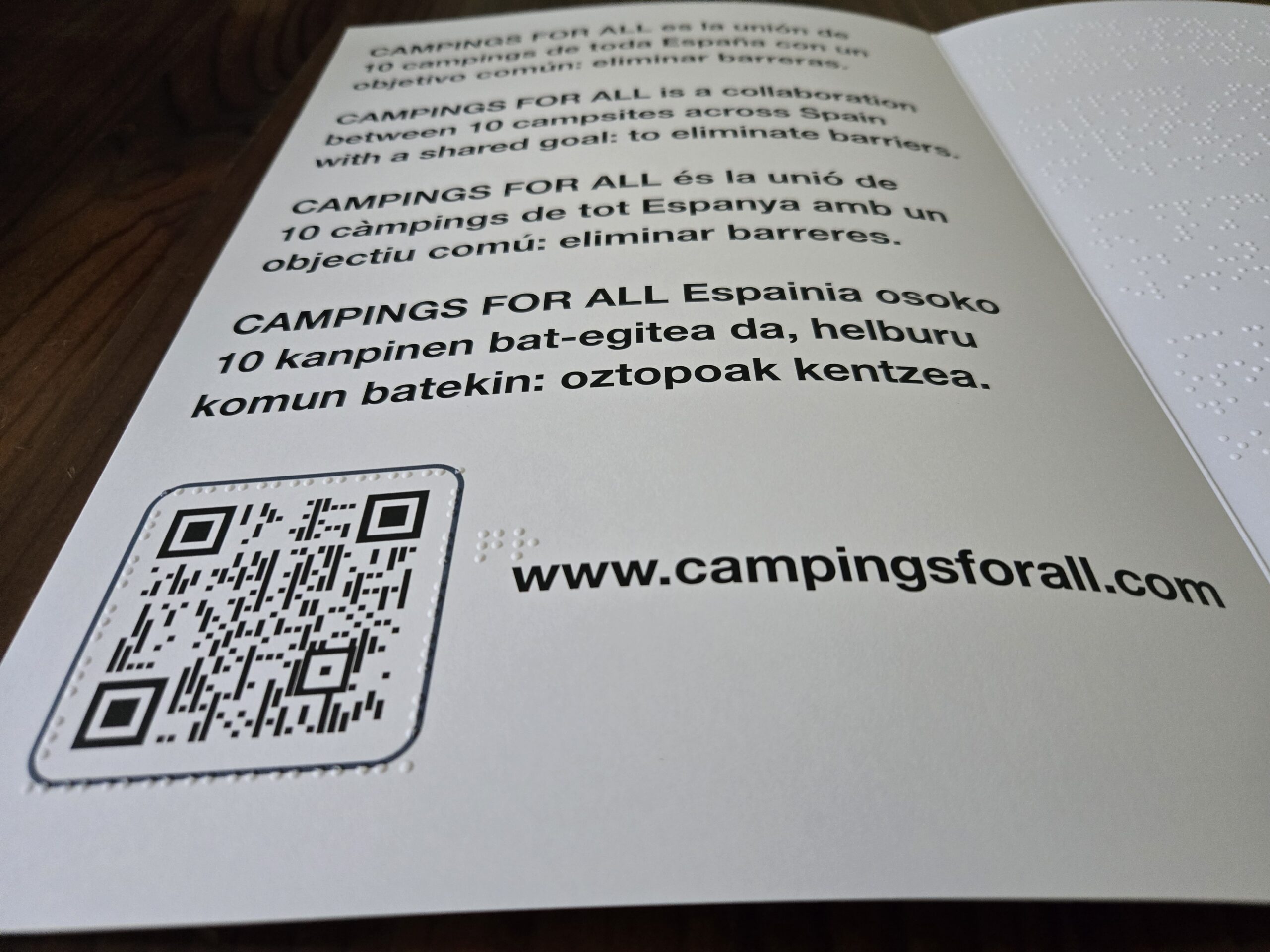 Doble pàgina oberta. A l'esquerra, text en macrocaràcters en quatre idiomes que explica que Campings For All agrupa deu càmpings d'Espanya per eliminar barreres, amb un codi QR i la URL campingsforall.com. A la dreta, el mateix text en braille en relleu.