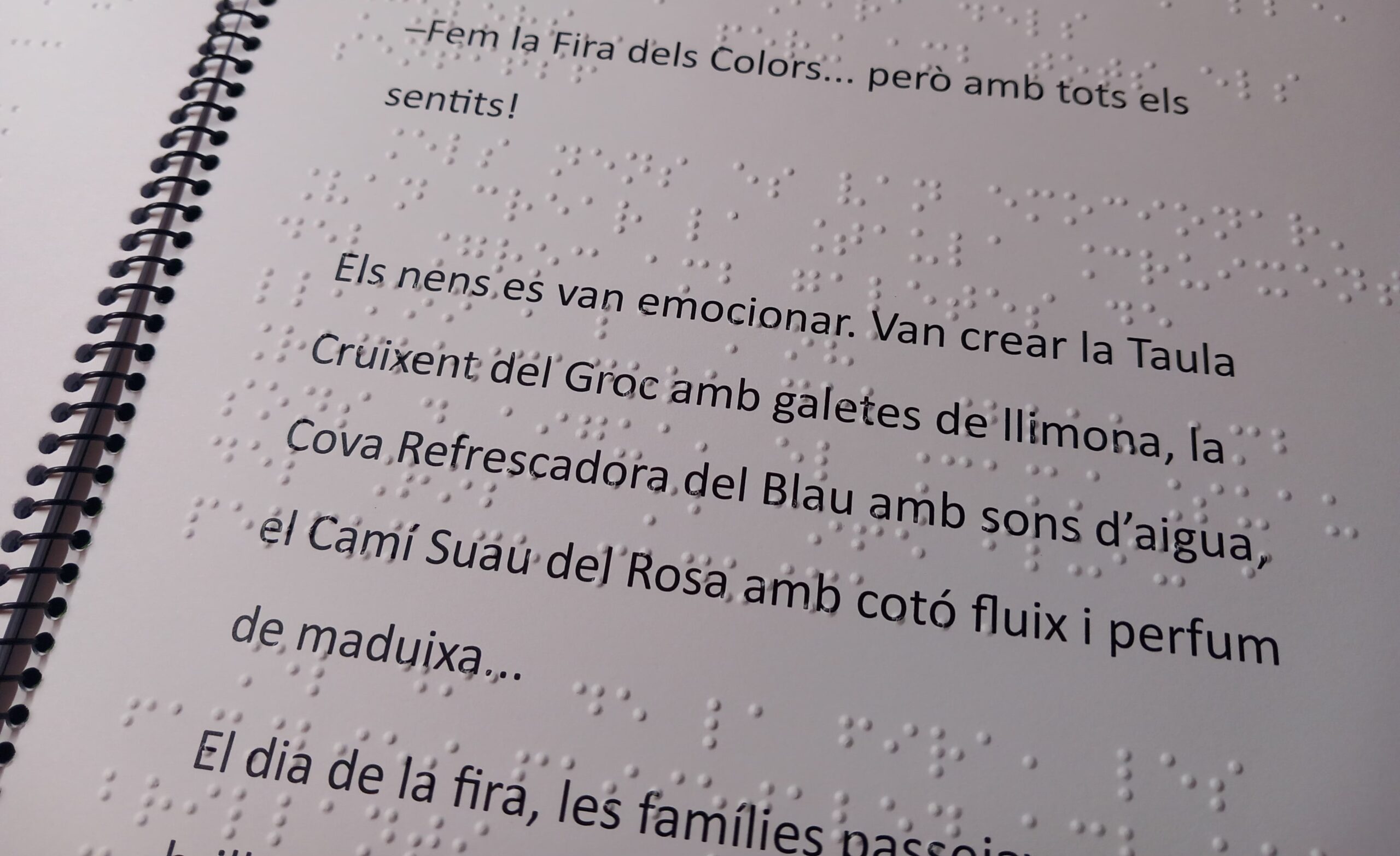 Pàgina interior del conte en macrocaràcters i braille. El text en tinta en lletra gran diu: "–Fem la Fira dels Colors... però amb tots els sentits! Els nens es van emocionar. Van crear la Taula Cruixent del Groc amb galetes de llimona, la Cova Refrescadora del Blau amb sons d'aigua, el Camí Suau del Rosa amb cotó fluix i perfum de maduixa...". Per sobre, hi ha escriptura braille en relleu.