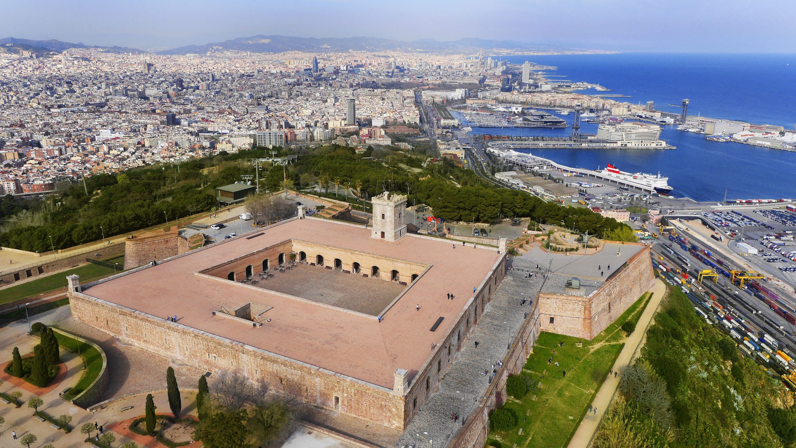 Vista aèria d’una fortalesa de pedra situada sobre una muntanya amb la ciutat i el port de Barcelona al fons.