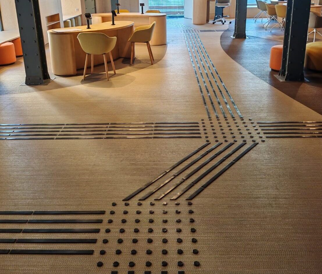 Sala àmplia amb mobiliari modern en tons càlids. Al terra, franges i botons negres formen el recorregut podotàctil que travessa l'espai central.